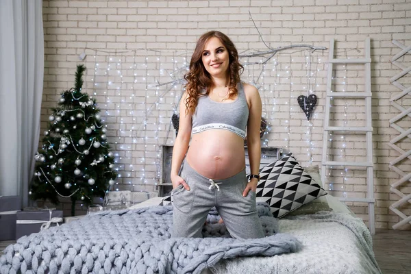 ランジェリーの妊娠中のブルネットが彼女の胃 幸せな母親を見てベッドの上に座っています 大きな灰色のベッドと灰色の柔らかいアームチェア おもちゃとクリスマスツリー 新年の雰囲気 — ストック写真