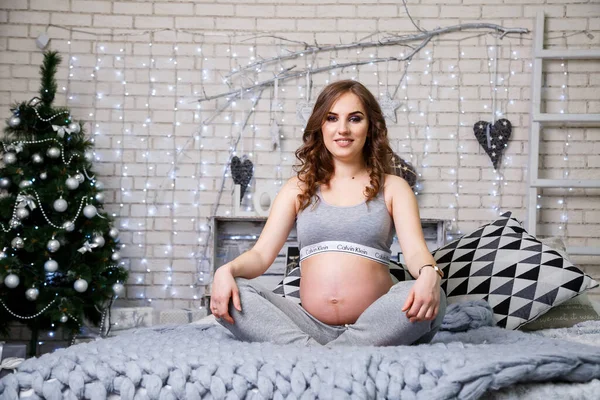 İç çamaşırlı hamile bir esmer yatakta oturmuş karnına bakıyor, mutlu annelik. Büyük gri yatak ve gri yumuşak koltuklar. Oyuncaklı Noel ağacı, Yeni Yıl atmosferi