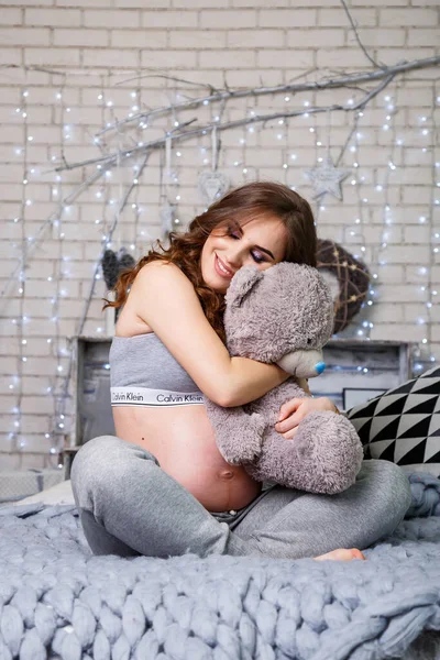 ランジェリーの妊娠中のブルネットは 彼女の胃 幸せな母親を見て ベッドの上に柔らかいクマと座っています 大きな灰色のベッド — ストック写真