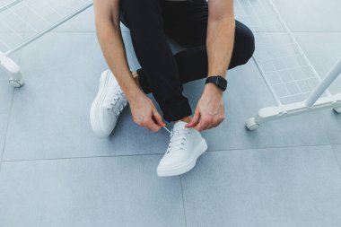 Beyaz derili, dantelli spor ayakkabılı ve siyah pantolonlu erkekler gri fayanslı arka planda.