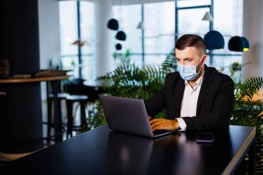 Coronavirus salgını sırasında bir kafede dizüstü bilgisayarla çalışan maskeli yakışıklı bir genç..