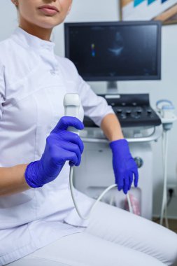 Genç bir kadın stajyer doktor, elinde ultrason cihazı tutan beyaz bir takım elbise ve tıbbi eldiven giyiyor. Ultrason kontrolleri. seçici odak