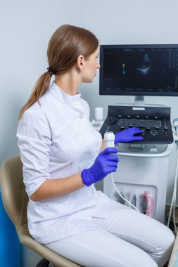 Genç bir kadın stajyer doktor, elinde ultrason cihazı tutan beyaz bir takım elbise ve tıbbi eldiven giyiyor. Ultrason kontrolleri. seçici odak