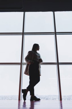 Pantolon ve ceket giyen başarılı bir iş kadını iş merkezi gökdeleninin devasa panoramik penceresinden dışarı bakar..