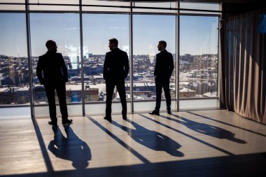 Panoramik pencerenin arka planında koyu renk erkek siluetleri var. Erkek işadamları metropol manzaralı bir gökdelenin geniş penceresinden dışarı bakıyor.