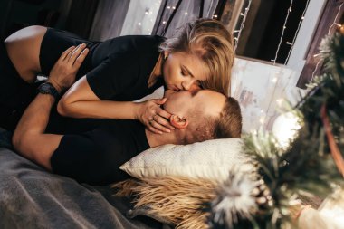 Yeni yıldan önce evde çelenklerle vakit geçiren tutkulu romantik çift manzarası. Sarışın bir genç kadın ve yakışıklı bir adam yatakta uzanıyorlar ve sarılıyorlar, öpüşüyorlar. Noel zamanı.