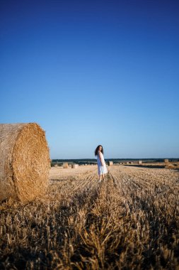 Beyaz bir yaz elbisesi içinde genç ve güzel bir kadın biçilmiş buğday tarlasında duruyor. Kocaman saman demetleriyle, doğanın tadını çıkarıyor. Köyün doğası. Seçici odak