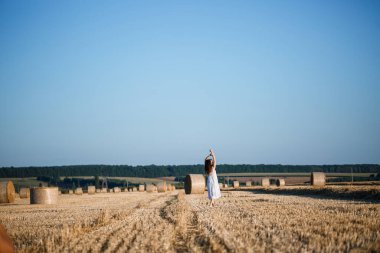 Beyaz bir yaz elbisesi içinde genç ve güzel bir kadın biçilmiş buğday tarlasında duruyor. Kocaman saman demetleriyle, doğanın tadını çıkarıyor. Köyün doğası. Seçici odak