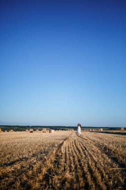Beyaz bir yaz elbisesi içinde genç ve güzel bir kadın biçilmiş buğday tarlasında duruyor. Kocaman saman demetleriyle, doğanın tadını çıkarıyor. Köyün doğası. Seçici odak