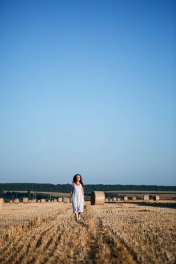 Beyaz bir yaz elbisesi içinde genç ve güzel bir kadın biçilmiş buğday tarlasında duruyor. Kocaman saman demetleriyle, doğanın tadını çıkarıyor. Köyün doğası. Seçici odak