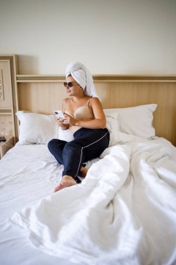 Güzel bir genç kadın başının üstünde havluyla büyük bir kar beyazı yatakta yatıyor..