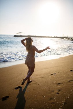 Güneş gözlüklü, bej bluzlu genç bir kadın gün batımında deniz kıyısında yürüyor.