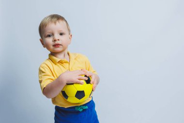 Elinde beyaz bir arka planda futbol topu olan küçük bir çocuk, acemi bir futbolcu, çocuklar için bir spor. Küçük atlet. Çocuklar için sarı ve mavi futbol takımı