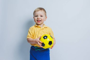 Elinde beyaz bir arka planda futbol topu olan küçük bir çocuk, acemi bir futbolcu, çocuklar için bir spor. Küçük atlet. Çocuklar için sarı ve mavi futbol takımı