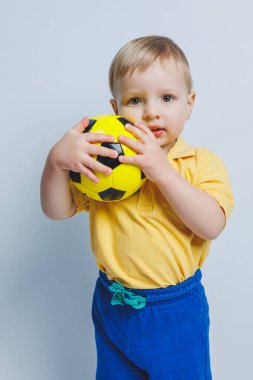 Elinde beyaz bir arka planda futbol topu olan küçük bir çocuk, acemi bir futbolcu, çocuklar için bir spor. Küçük atlet. Çocuklar için sarı ve mavi futbol takımı