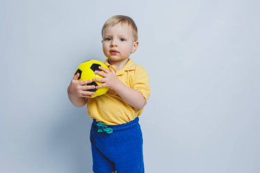Elinde beyaz bir arka planda futbol topu olan küçük bir çocuk, acemi bir futbolcu, çocuklar için bir spor. Küçük atlet. Çocuklar için sarı ve mavi futbol takımı