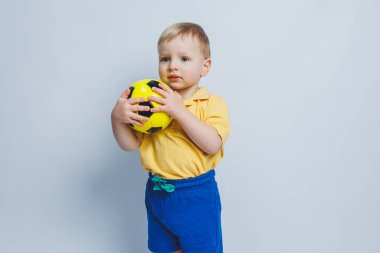 Elinde futbol topu olan sarı tişörtlü küçük bir çocuk beyaz arka planda izole edilmiş bir şekilde gülümsüyor. Spor çocuğu elinde top tutuyor. Çocuk sporları oyunu. Küçük sporcu..