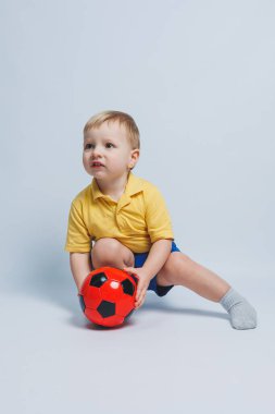 3-4 yaşlarında, sarı tişörtlü bir futbol fanatiği elinde bir top, beyaz bir arka planda izole bir futbol topu tutuyor. Spor aile eğlencesi konsepti