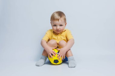 3-4 yaşlarında, sarı tişörtlü bir futbol fanatiği elinde bir top, beyaz bir arka planda izole bir futbol topu tutuyor. Spor aile eğlencesi konsepti