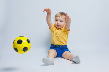 5-6 yaşlarında sarı tişörtlü neşeli bir çocuk, takımı destekliyor, elinde bir futbol topu tutuyor, beyaz bir arka planda izole edilmiş. Spor aile eğlencesi konsepti