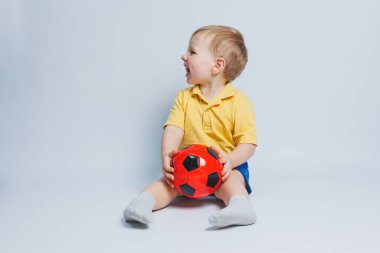 Elinde futbol topu olan sarı tişörtlü küçük bir çocuk beyaz arka planda izole edilmiş bir şekilde gülümsüyor. Spor çocuğu elinde top tutuyor. Çocuk sporları oyunu. Küçük sporcu..