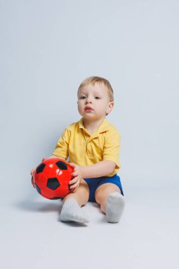 5-6 yaşlarında sarı tişörtlü neşeli bir çocuk, takımı destekliyor, elinde bir futbol topu tutuyor, beyaz bir arka planda izole edilmiş. Spor aile eğlencesi konsepti