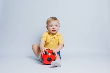 3-4 yaşlarında, sarı tişörtlü bir futbol fanatiği elinde bir top, beyaz bir arka planda izole bir futbol topu tutuyor. Spor aile eğlencesi konsepti