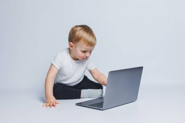 3-4 yaşlarında küçük bir çocuk beyaz arka planda dizüstü bilgisayarla oturuyor. Beyaz tişörtlü ve siyah pantolonlu bir çocuk dizüstü bilgisayara bakıyor. Modern çocuklar