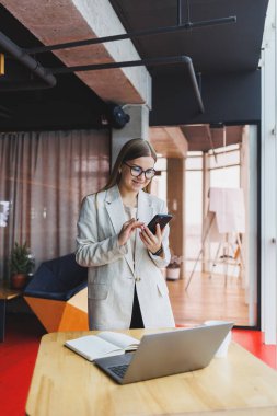 Modern çalışma ortamında açık bir ağ kitabıyla oturan ve günlük kıyafetlerle cep telefonundan bildirileri kontrol eden kadın serbest çalışan..