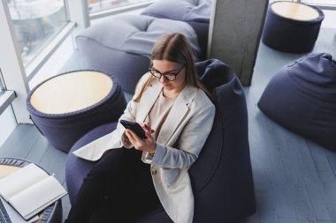 Modern çalışma ortamında açık bir ağ kitabıyla oturan ve günlük kıyafetlerle cep telefonundan bildirileri kontrol eden kadın serbest çalışan..