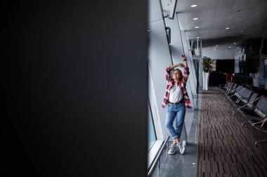 Sarışın, güzel bir Ukraynalı kadın geniş bir pencerenin yanında duruyor, momom kot pantolon ve ekose gömlekli bir tişört giyiyor. Rahat giyimli kız.