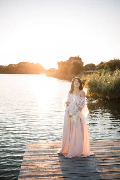 一位身穿雪纺长裙的年轻孕妇站在河边的码头上 背景是橙色的日落 — 图库照片