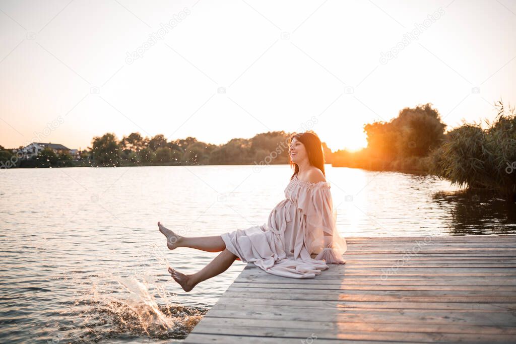 A young pregnant woman in a delicate dress by the river with a beautiful sunset. Beauty, health and gentle motherhood. happy motherhood