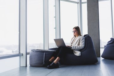 Başarılı, ceketli, gülümseyen ve geniş pencereli, modern ofiste dizüstü bilgisayarla çalışan genç bir kadın. Uzak çalışma