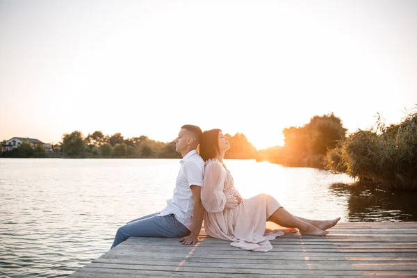 一个丈夫和一个怀孕的妻子正在河岸上休息 在夏日落日的晴天坐在码头上 恋爱中的男女手牵手拥抱 — 图库照片