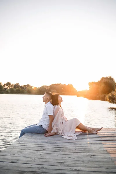 一个丈夫和一个怀孕的妻子正在河岸上休息 在夏日落日的晴天坐在码头上 恋爱中的男女手牵手拥抱 — 图库照片