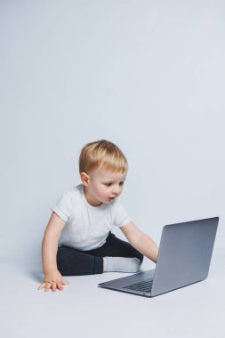 3-4 yaşlarında küçük bir çocuk beyaz arka planda dizüstü bilgisayarla oturuyor. Beyaz tişörtlü ve siyah pantolonlu bir çocuk dizüstü bilgisayara bakıyor. Modern çocuklar
