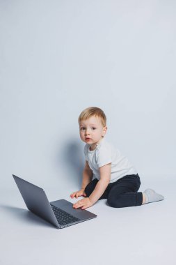 Akıllı bir çocuk 3-4 yaşlarında beyaz bir arka planda dizüstü bilgisayarla oturuyor. Beyaz tişörtlü ve siyah pantolonlu bir çocuk dizüstü bilgisayarın başına oturur ve kameraya bakar. Modern çocuklar