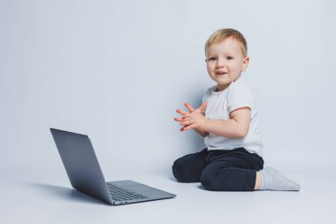 Akıllı bir çocuk 3-4 yaşlarında beyaz bir arka planda dizüstü bilgisayarla oturuyor. Beyaz tişörtlü ve siyah pantolonlu bir çocuk dizüstü bilgisayarın başına oturur ve kameraya bakar. Modern çocuklar