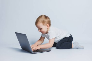 3-4 yaşlarında küçük zeki bir çocuk beyaz bir arka planda dizüstü bilgisayarla oturuyor. Beyaz tişörtlü ve siyah pantolonlu bir çocuk dizüstü bilgisayarın başına oturur ve ekrana bakar. Modern yenilikçi çocuklar
