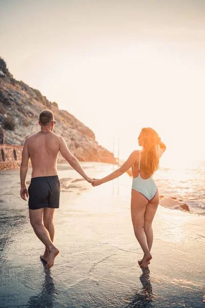 愛するカップルが海でビーチを歩いています 地中海に沈む夕日の若い家族 休暇の概念 水着姿の女性と海に沈む夕日のショートパンツ姿の男性 選択的焦点 — ストック写真