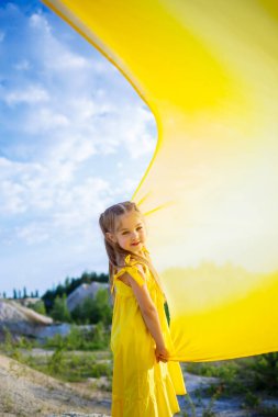 Göl kenarında sarı kumaşlı sarı kanatlı sarı elbiseli 6-7 yaşlarında Ukraynalı bir kız. Mavi gökyüzü ve sarı kumaş, Ukrayna bayrağının renkleri..