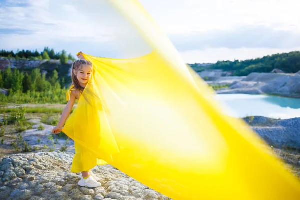 Göl kenarında sarı kumaşlı sarı kanatlı sarı elbiseli 6-7 yaşlarında Ukraynalı bir kız. Mavi gökyüzü ve sarı kumaş, Ukrayna bayrağının renkleri..