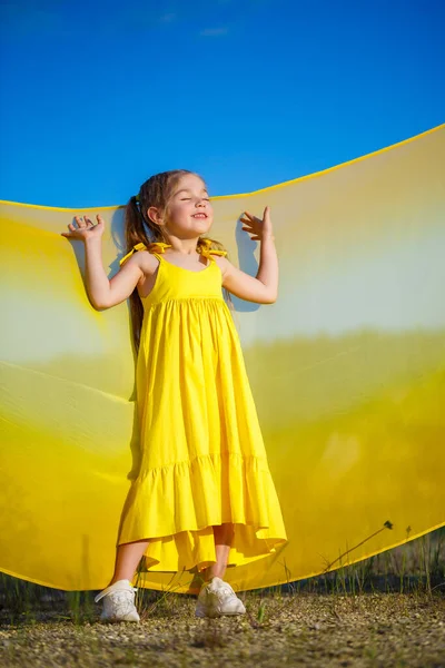 Ukraynalı 7 yaşında, Ukraynalı bir vatansever, mavi gökyüzüne karşı sarı bir elbise giymiş. Mavi gökyüzü. Güneşli bir gün. Özgür Ukraynalı çocuk. Ukrayna 'nın çocuk sembolü