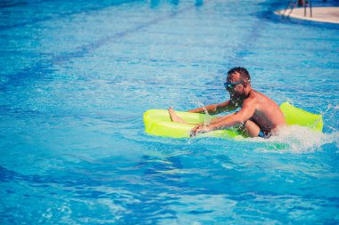 Yakışıklı genç adam mavi havuzda şişme yatakta yüzüyor.