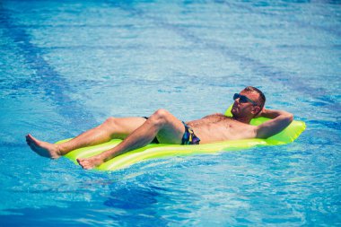 Genç bir adam havuzda sarı şişme bir şiltenin üzerinde banyo yapıyor. Tatilde güneşli güneşte rahatla. Mutlu başarılı adam.