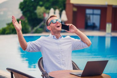 Genç erkek iş adamı, dizüstü bilgisayarı havuz kenarında oturuyor. Açık işyeri. Serbest yazar konsepti. İnternetten alışveriş. Tatil işi.