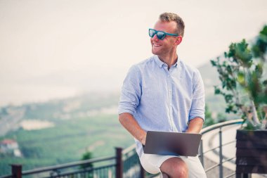 Genç başarılı erkek işadamı deniz kenarında dizüstü bilgisayarla çalışıyor. Güneş gözlüğü, gömlek ve beyaz şort giyiyor. Ofisin dışında çalış, serbest çalışan.