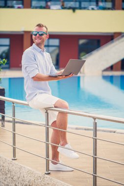 Genç erkek iş adamı, dizüstü bilgisayarı havuz kenarında oturuyor. Açık işyeri. Serbest yazar konsepti. İnternetten alışveriş. Tatil işi.