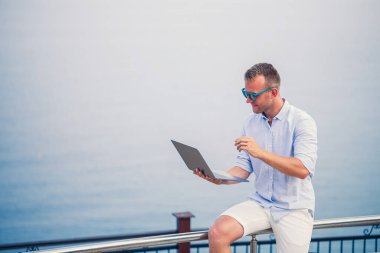 Genç başarılı erkek işadamı deniz kenarında dizüstü bilgisayarla çalışıyor. Güneş gözlüğü, gömlek ve beyaz şort giyiyor. Ofisin dışında çalış, serbest çalışan.